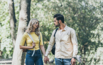 A man and woman walk hand-in-hand through a forested area. The woman wears a yellow top and jeans, carrying a backpack over her shoulder. The man wears a beige shirt and jeans, also carrying a backpack. Both look at each other as they walk along the wooded path.