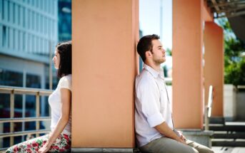 A woman and a man sit back-to-back against a large pillar outdoors. The woman, wearing a white shirt and floral pants, faces left, while the man, in a white shirt and beige pants, faces right. The setting appears urban, with buildings and railings in the background.
