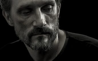 A close-up, black-and-white photograph of a middle-aged man with a beard and short hair. He gazes downward with a somber expression, highlighting the texture of his skin and the deep shadows on his face. He wears a dark shirt against a black background.