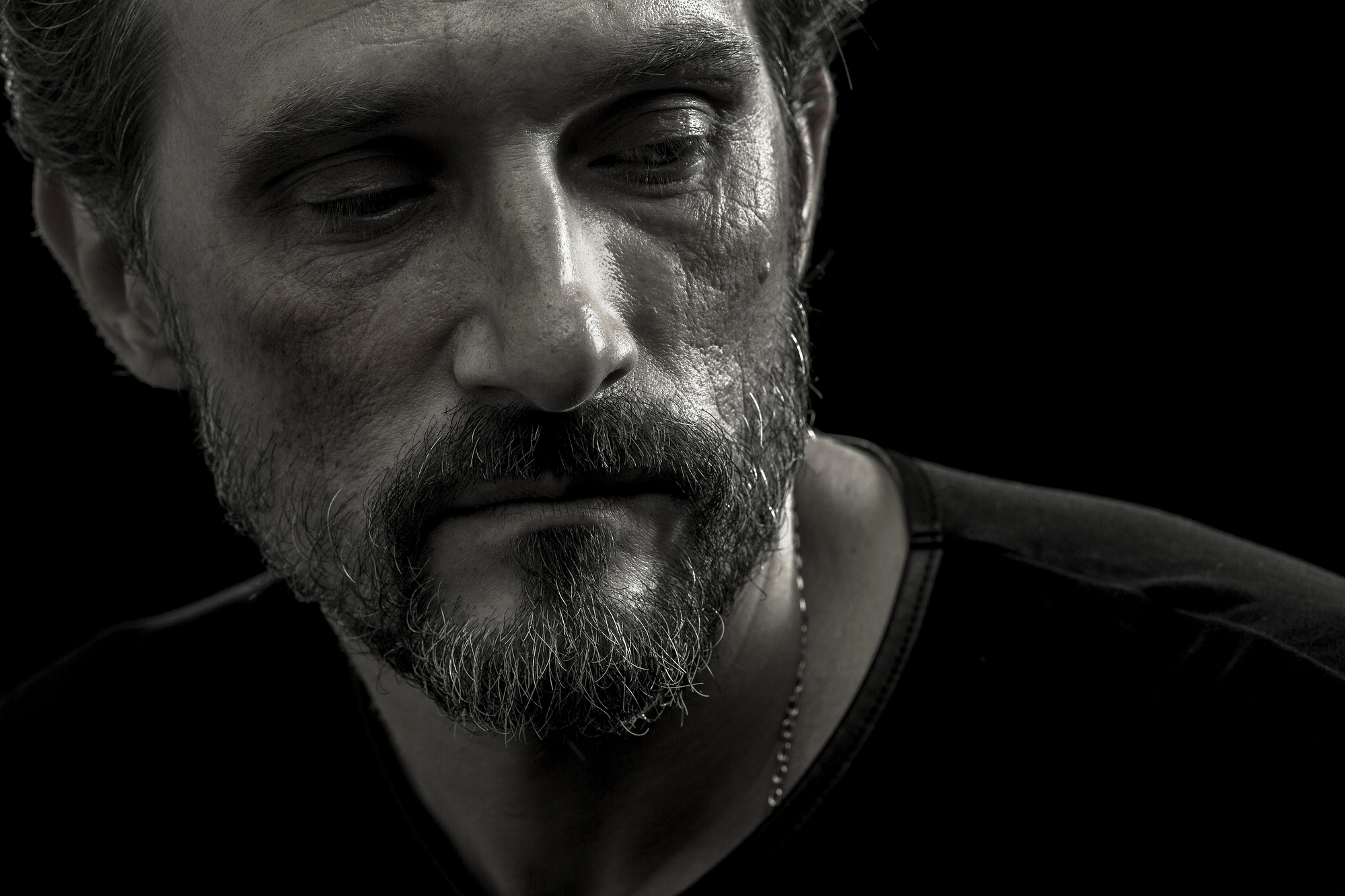 A close-up, black-and-white photograph of a middle-aged man with a beard and short hair. He gazes downward with a somber expression, highlighting the texture of his skin and the deep shadows on his face. He wears a dark shirt against a black background.