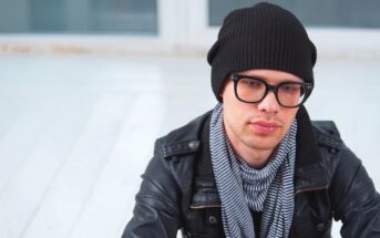 A person with glasses, wearing a black beanie, black leather jacket, and a striped scarf, is sitting in an outdoor setting with a light-colored wooden floor and blurred background. The person appears to be looking off to the side with a neutral expression.