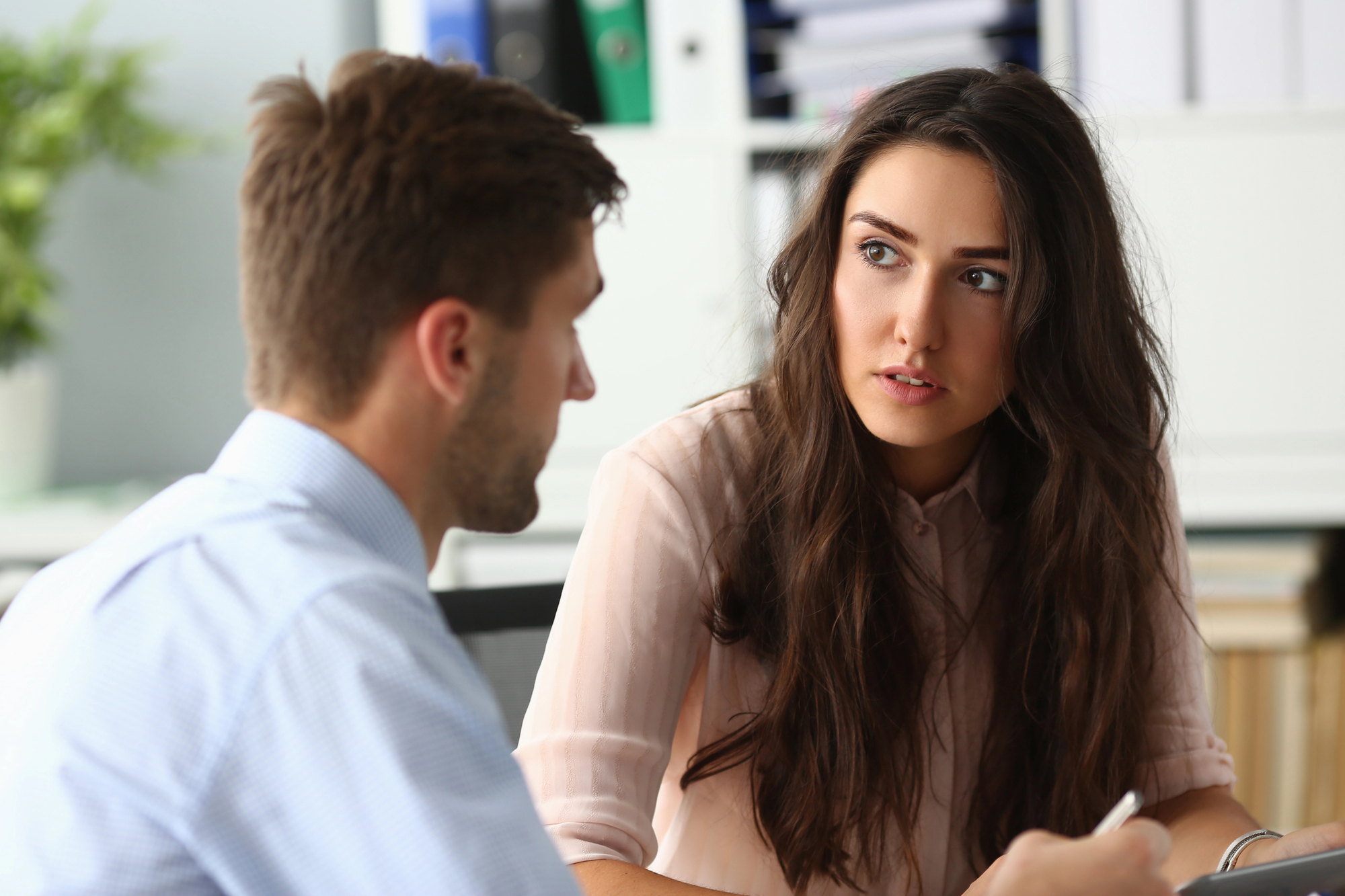 A man and a woman are sitting at a desk in an office setting, engaged in a conversation. The woman is looking attentively at the man, who has his back to the camera. Shelves with binders and books are visible in the background.