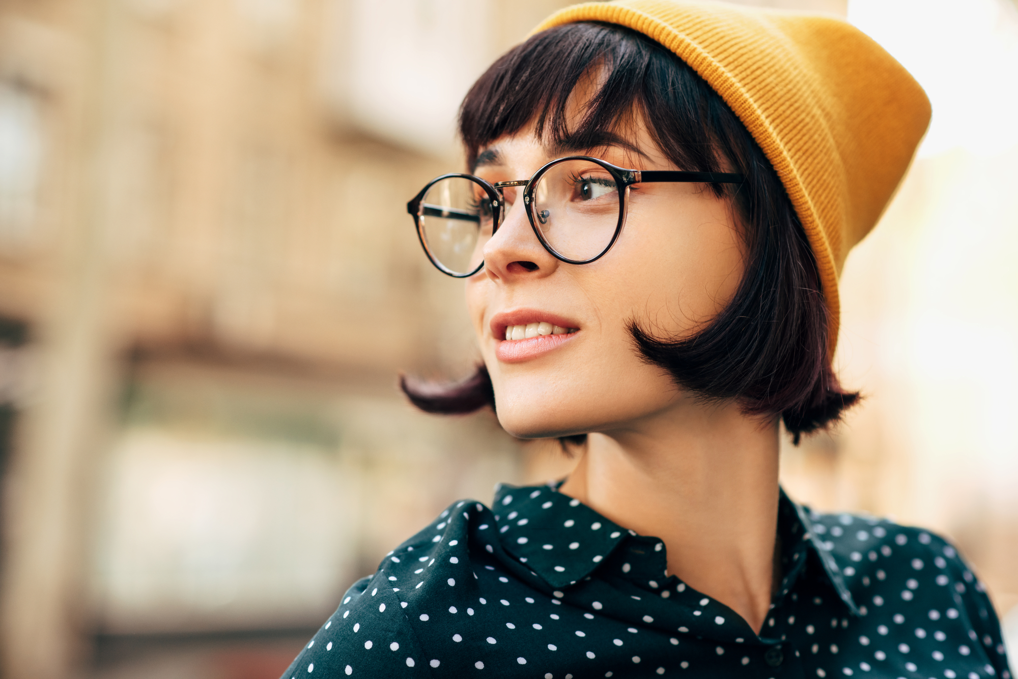 A young woman with short dark hair, wearing large round glasses, a yellow beanie, and a dark green polka dot shirt, smiles while looking off to the side. The background is blurred cityscape.