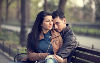 A young couple sits on a park bench; the woman, with long dark hair, wears a brown jacket and blue shirt, gazing to the side. The man, with short dark hair, wears a dark jacket, embracing her from behind and resting his chin on her shoulder, looking forward.