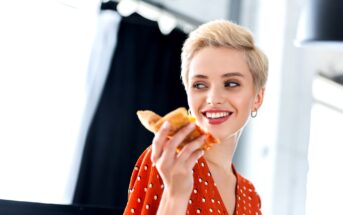 A person with short blonde hair is smiling and looking to the side while holding a slice of pizza. They are wearing a red polka dot top and silver hoop earrings. The background is blurred, but it appears to be indoors with natural light coming through a window.