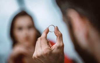 A close-up of a person holding a ring in their hand, with the ring clearly in focus. In the blurred background, another person is looking at the ring. The scene suggests an emotional or significant moment between the two individuals.