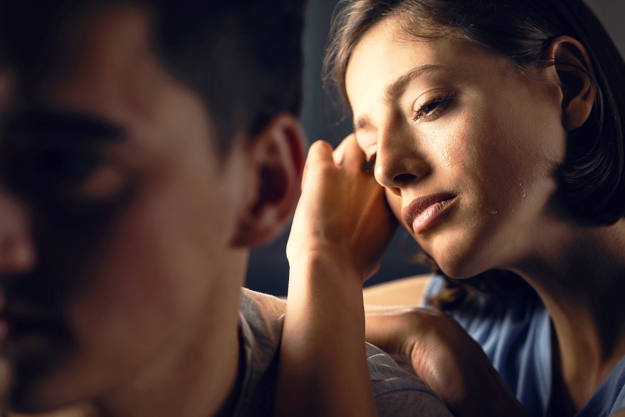 A woman with a pensive expression rests her head on her hand, looking closely at a man who is facing away from the camera, out of focus. The image conveys a sense of contemplation and emotion shared between the two individuals.