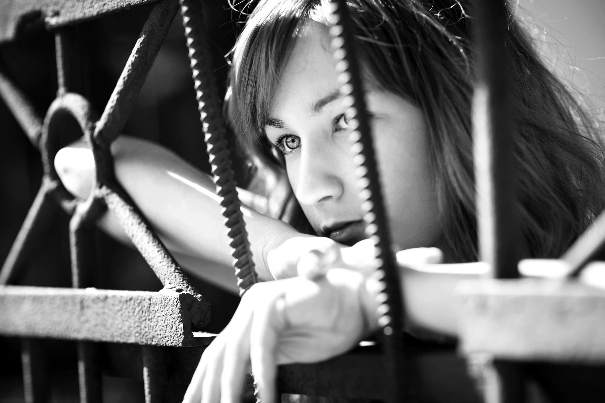 Black and white close-up of a woman with long hair, resting her arms and head on a rusted metal fence. She gazes thoughtfully into the distance, with sunlight casting shadows across her face and the fence.