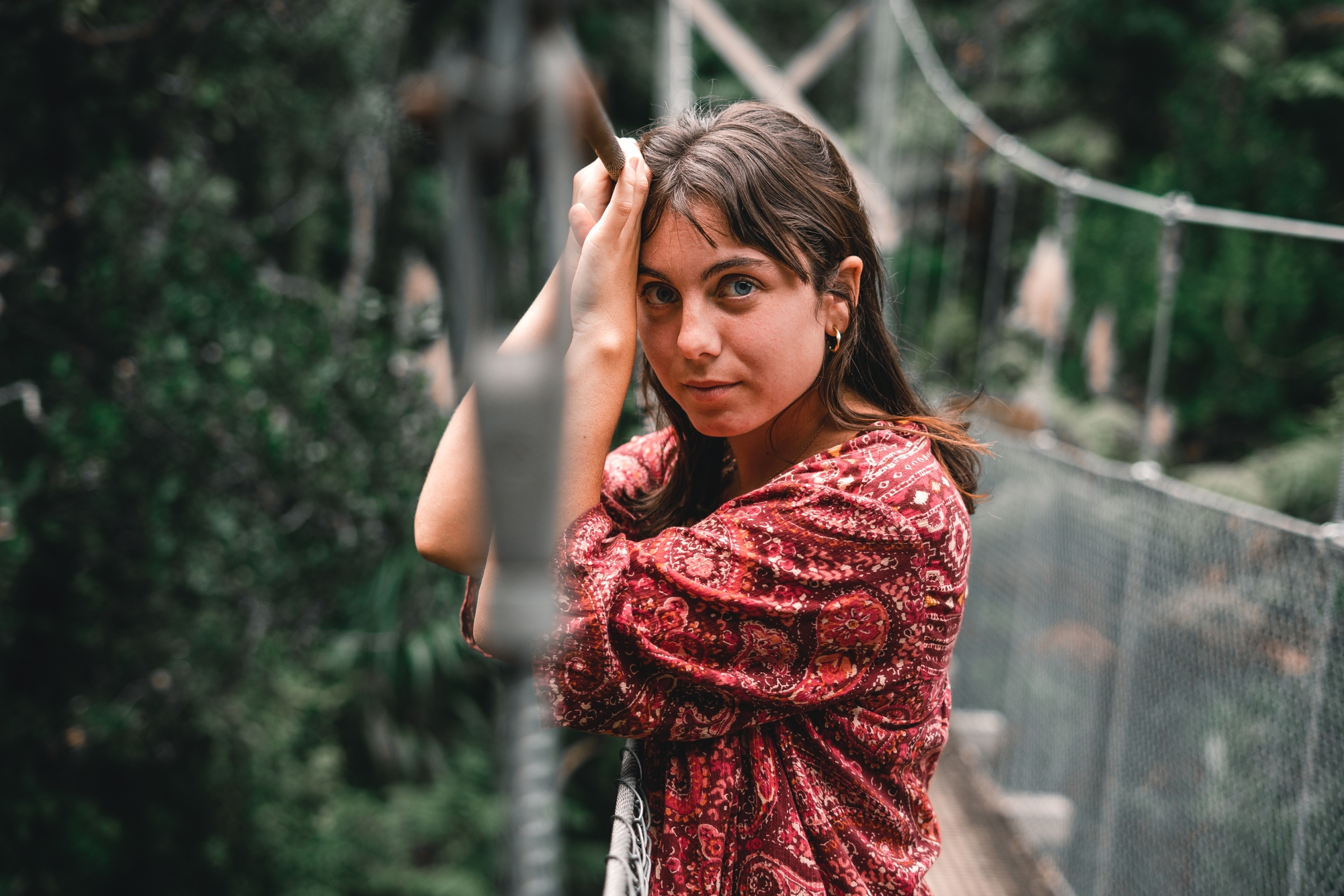 A woman with long brown hair is standing on a suspension bridge in a lush green forest. She is leaning on the bridge ropes with her hand resting on her head. She is wearing a red-patterned dress and looking directly at the camera with a serious expression.