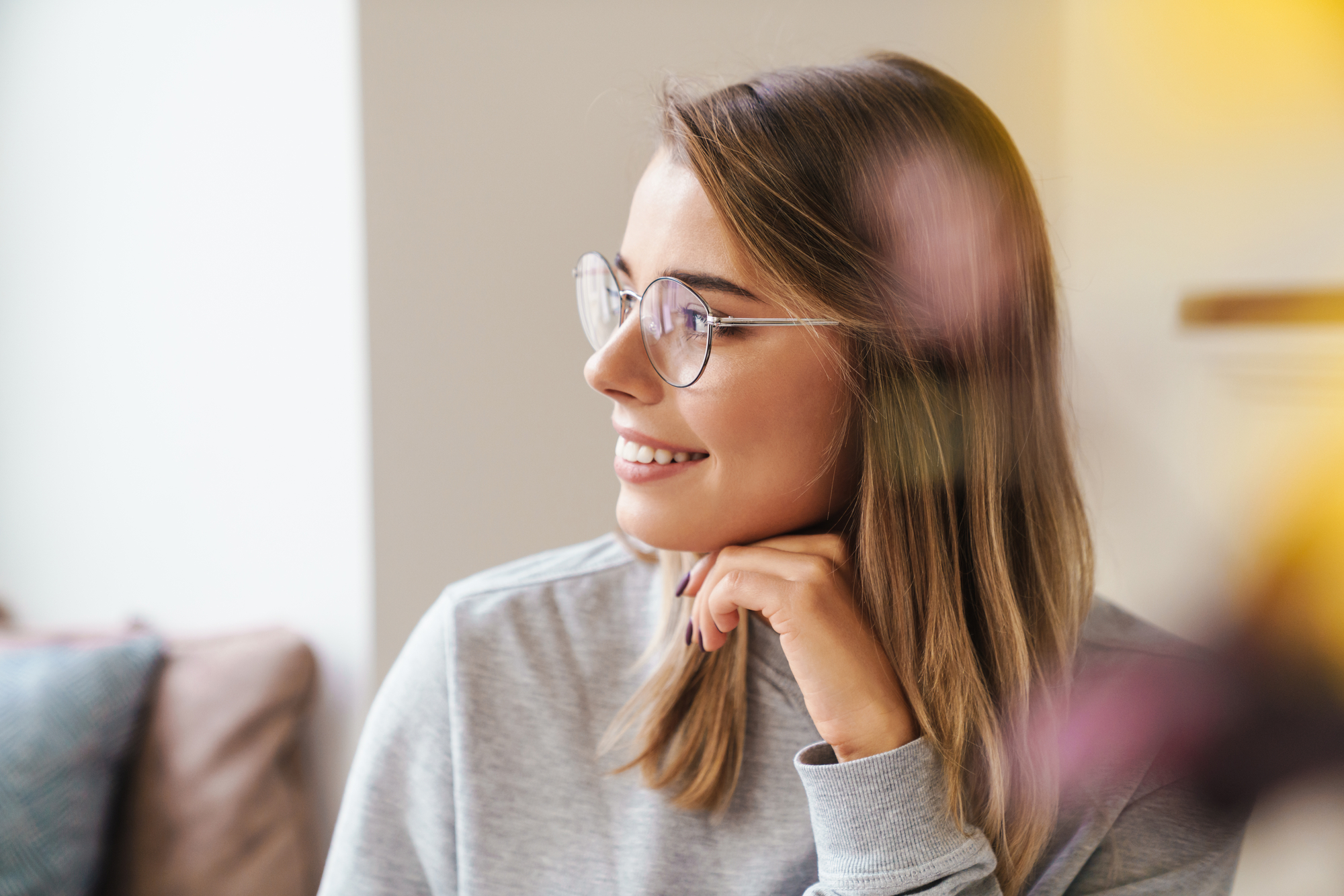 A woman with shoulder-length blond hair and glasses is smiling while looking to the side. She has her hand resting under her chin. She is indoors, with a blurred background that includes soft tones of yellow and pink.