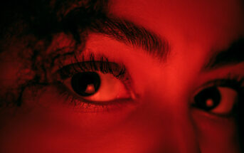 Close-up image of a person's eyes and eyebrows illuminated by red light. The focus is on the eyes, which are looking directly at the camera. Strands of curly hair are partially visible on the left side of the image.
