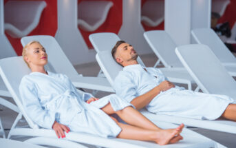A woman and a man in white bathrobes are reclining on white lounge chairs in a spa, appearing relaxed. The background features modern white and red decor and additional empty lounge chairs.