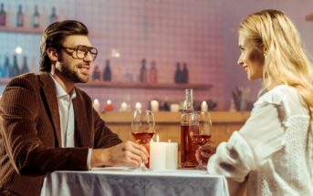 A man and a woman are seated at a candlelit table in a dimly lit restaurant, enjoying glasses of rosé wine. The man is dressed in a brown suit jacket and glasses, while the woman is in a white blouse. Both are smiling and engaged in conversation.