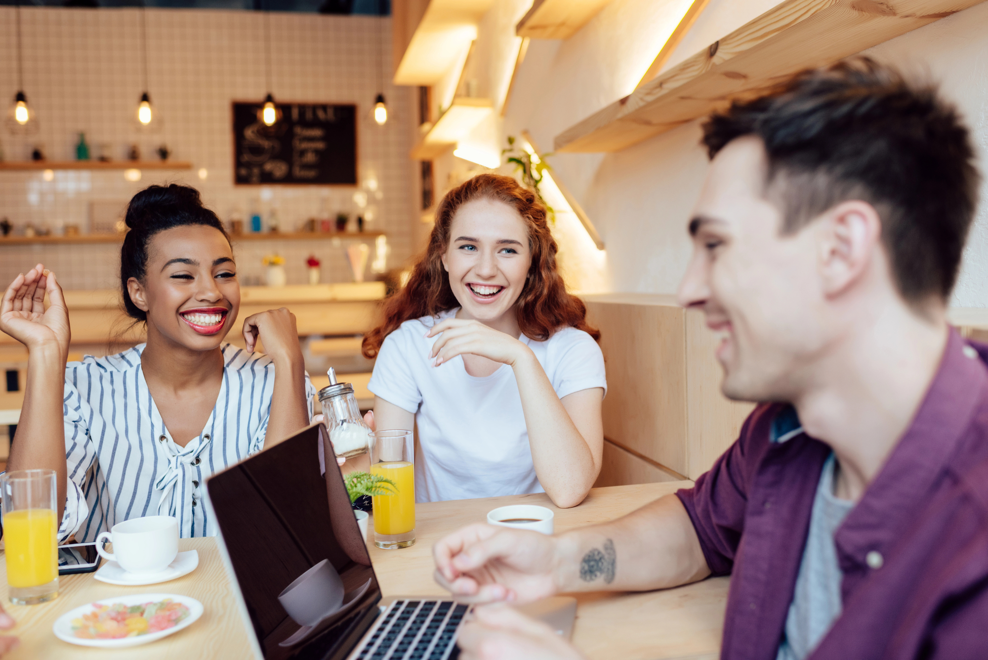 Three people are seated around a table in a casual café setting, smiling and engaging in conversation. One person is using a laptop. Drinks and a plate of food are on the table. The background shows a well-lit café interior with shelves and a menu board.