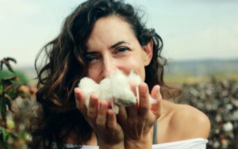 A woman with wavy brown hair holds fluffy white cotton close to her face, smiling gently. She is outdoors in a field, with blurred cotton plants visible in the background, wearing a white off-the-shoulder top.