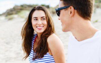 A woman with long brown hair, wearing a striped tank top, smiles at a man with short brown hair and sunglasses. They are sitting together on a sandy beach with blurred greenery in the background. The man is turned towards the woman, appearing to enjoy the moment.