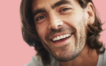 A man with long brown hair and a neatly trimmed beard is smiling warmly. He has a light complexion, and he's wearing a white top. The background is a soft pink hue, adding a pleasant contrast to the image. His expression conveys happiness and friendliness.