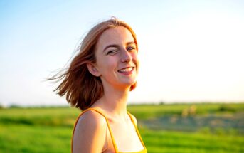 A young person with short, auburn hair smiles brightly while standing in a sunlit field. They are wearing an orange sleeveless top. The sky is clear and blue, and the surrounding grass is green and lush, creating a serene and cheerful atmosphere.