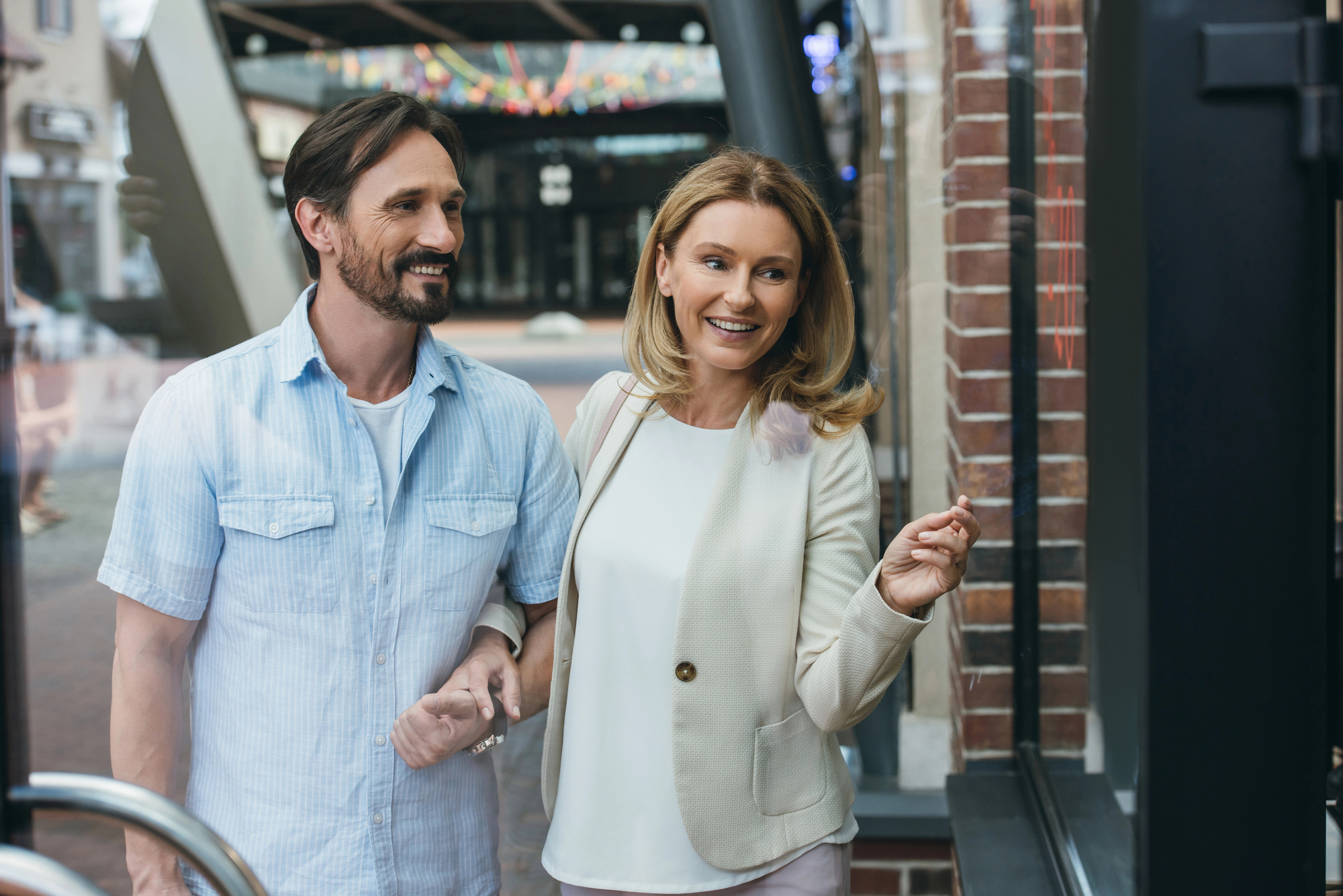 A smiling couple is standing outside a store, holding hands and looking through the window. The man has dark hair and is wearing a light blue shirt, while the woman has blonde hair and is wearing a white jacket. They appear happy and engaged in window shopping.