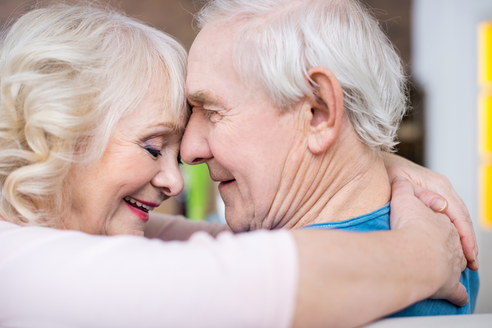 An elderly couple with white hair embraces, their foreheads touching, and they smile warmly at each other. The woman wears a light pink top, and the man is in a blue shirt. The image captures a tender and loving moment between them.