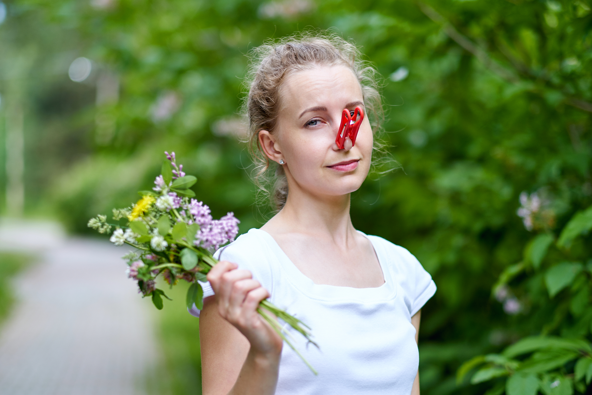 A woman with blonde hair is standing outdoors, holding a small bouquet of wildflowers. She is wearing a white shirt and has a red clothespin clipped to her nose. The background is lush with green foliage.