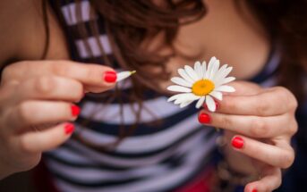 Close-up of a person with red-painted nails plucking petals from a daisy flower. The person, wearing a striped top, is holding the daisy in one hand while pulling a petal with the other. The background is blurred, focusing on the hands and daisy.