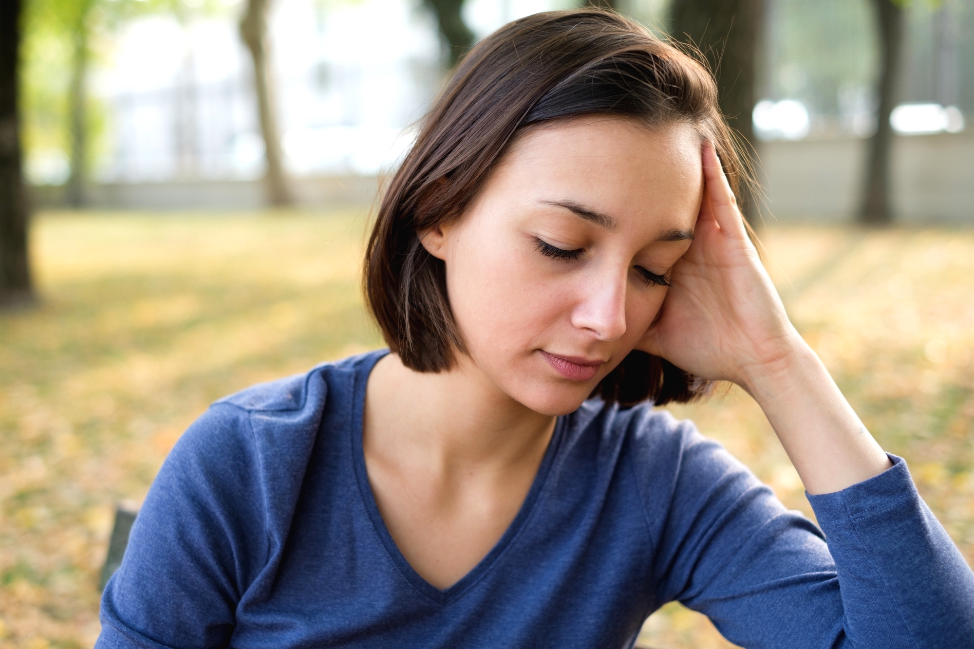 A young woman with short brown hair, wearing a blue long-sleeve shirt, rests her head on her hand while sitting outdoors in a park. She appears to be deep in thought, with her eyes closed and a neutral expression. The background is blurred with trees and fallen leaves.