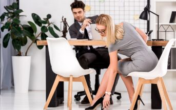 A woman in a gray dress bends down to pick up her black high heel shoe, which she dropped. She sits on a white chair in an office setting. A man in a suit sits across from her at a desk, looking at her. There's a large plant and office supplies in the background.