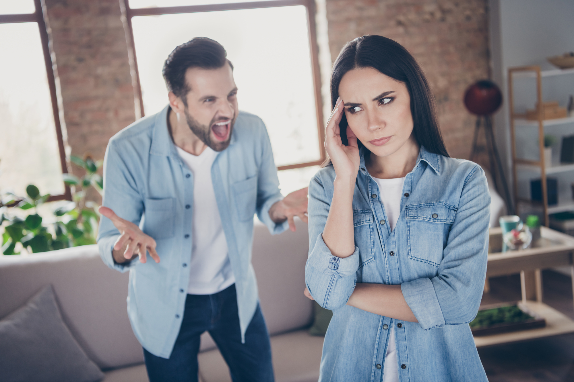 A man is angrily shouting at a woman in a living room with large windows. The woman, looking frustrated and upset, is standing with her arms crossed and her hand touching her temple. Both are wearing denim shirts. The room has shelves and a couch.