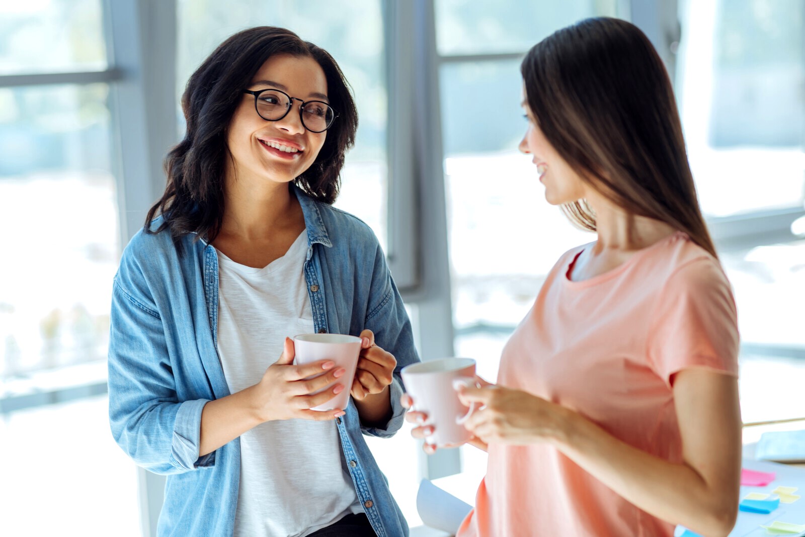 Two women are standing and holding white mugs, smiling at each other. The woman on the left is wearing glasses and a denim shirt, while the woman on the right has long hair and is wearing a peach-colored shirt. They appear to be in a bright, modern indoor setting.