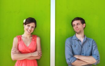 A woman in a pink dress with folded arms smiles while standing beside a man in a blue shirt, also with folded arms, who looks to the side with a thoughtful expression. They are in front of a bright green wall separated by a vertical white stripe.