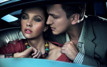 A woman with dark hair wearing a necklace and red top sits in a car, looking pensively out the window. A man in a gray suit leans towards her, gently touching her shoulder and collar, with an intense, intimate expression. Both have serious expressions.