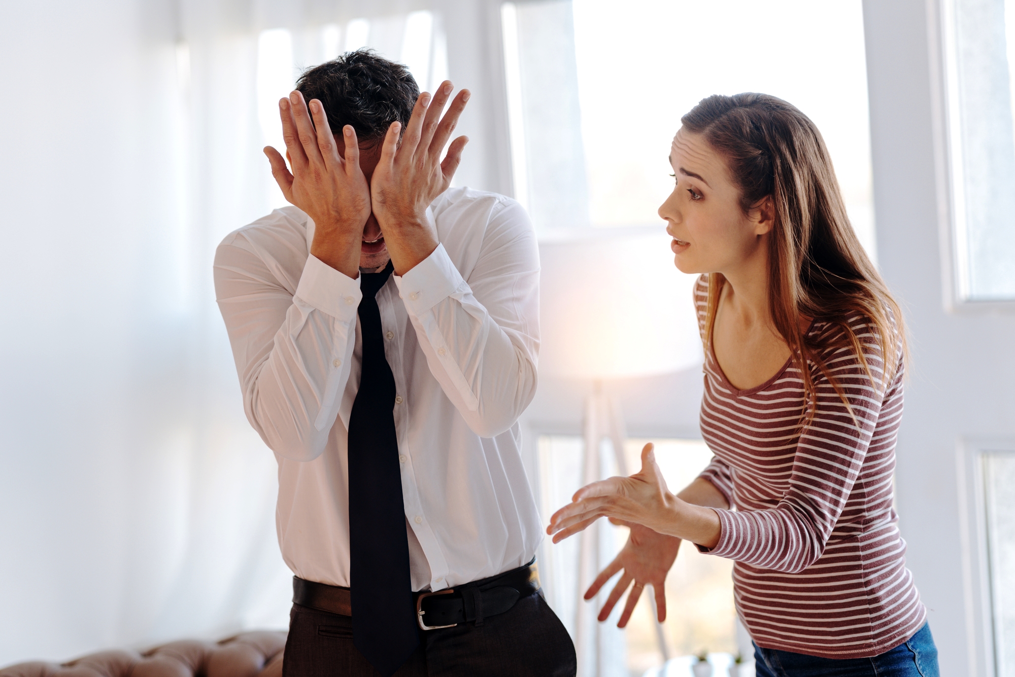 A woman with long brown hair, wearing a striped shirt, is gesturing and looking at a man in a white shirt and dark tie who is covering his face with his hands, appearing stressed or upset, in a bright room with natural light.