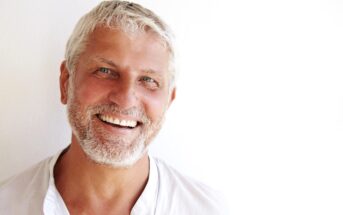 A smiling older man with short white hair and a beard stands against a plain white background. He is wearing a white shirt with an open collar. The lighting is bright, highlighting his features and creating a cheerful and warm atmosphere.