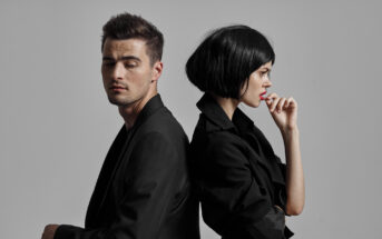 A man and a woman stand back-to-back against a gray background. Both are dressed in black. The man has his eyes closed and head slightly tilted to the side, while the woman, with a bob haircut, pensively touches her lip in profile view.