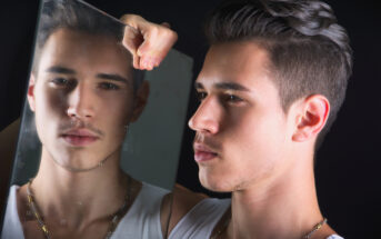 A young man with short dark hair and a light stubble is looking into a mirror, holding it with his right hand. His reflection in the mirror shows his pensive expression. He is wearing a white tank top and a gold chain necklace. The background is black.