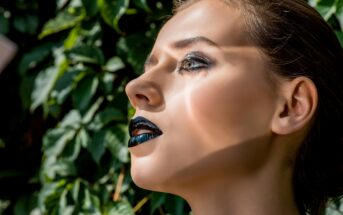 A close-up side profile of a woman with dramatic black and metallic makeup, including dark lipstick and bold eyeshadow. She has light skin and her hair pulled back, standing against a backdrop of green leaves illuminated by sunlight.