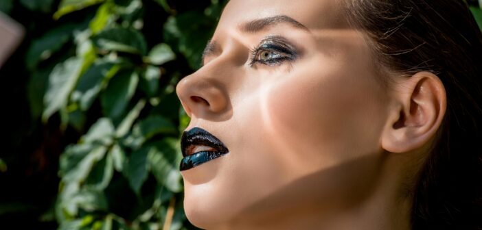 A close-up side profile of a woman with dramatic black and metallic makeup, including dark lipstick and bold eyeshadow. She has light skin and her hair pulled back, standing against a backdrop of green leaves illuminated by sunlight.