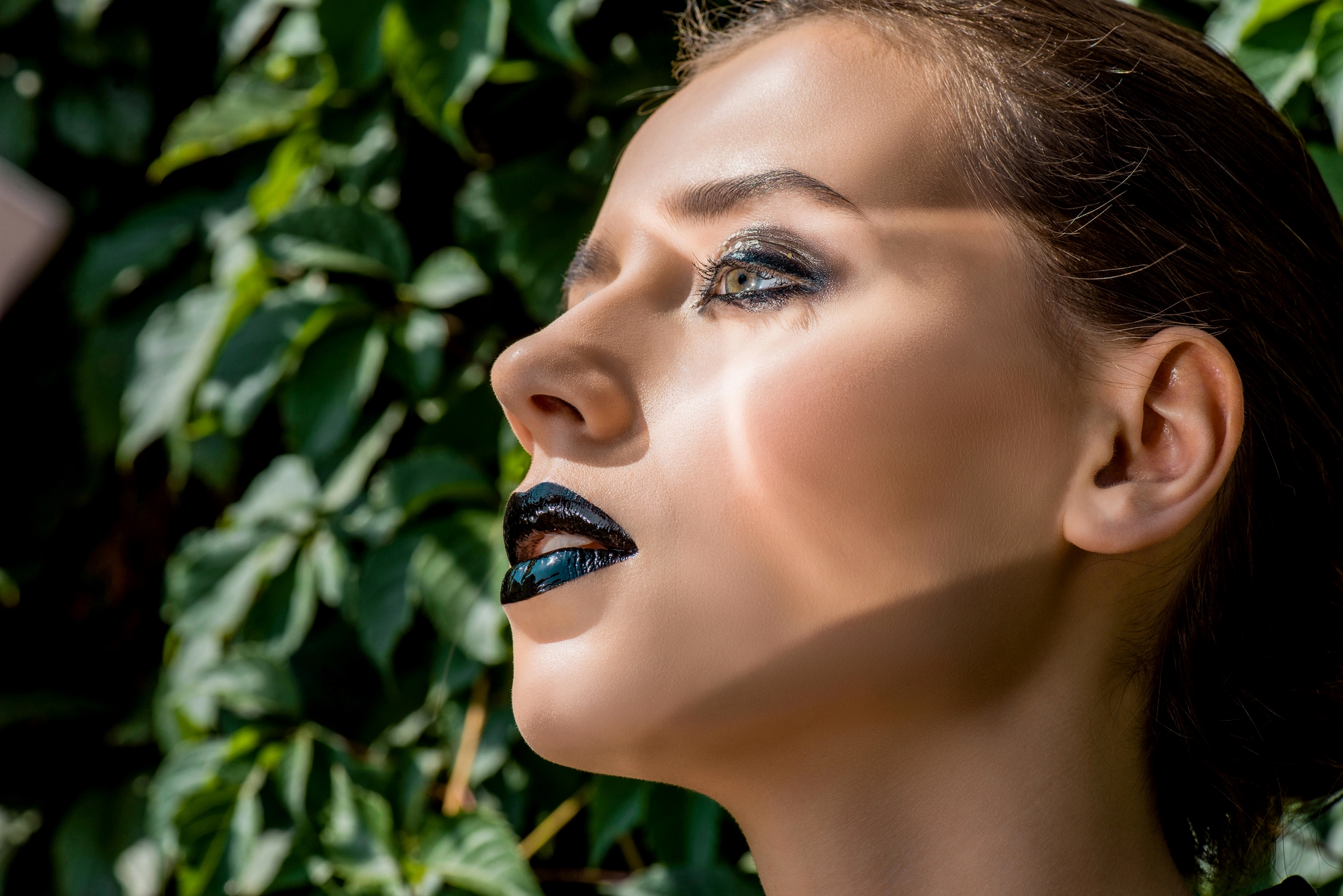 A close-up side profile of a woman with dramatic black and metallic makeup, including dark lipstick and bold eyeshadow. She has light skin and her hair pulled back, standing against a backdrop of green leaves illuminated by sunlight.