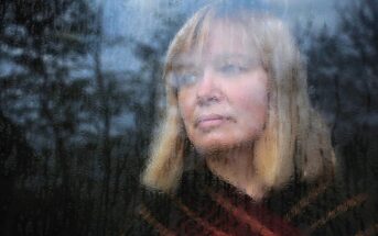 A woman with blonde hair looks pensively out of a foggy, rain-streaked window. The blurred glass obscures some details of her face and the dark, forest-like scenery outside. She wears a dark top with a red pattern. The mood appears contemplative and somber.