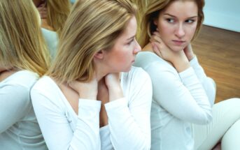 A woman with blonde hair, wearing a white long-sleeved shirt, is sitting in front of a mirror. She is looking at her reflection with a thoughtful expression, her hands clasped around her neck. Multiple reflections are visible around her.