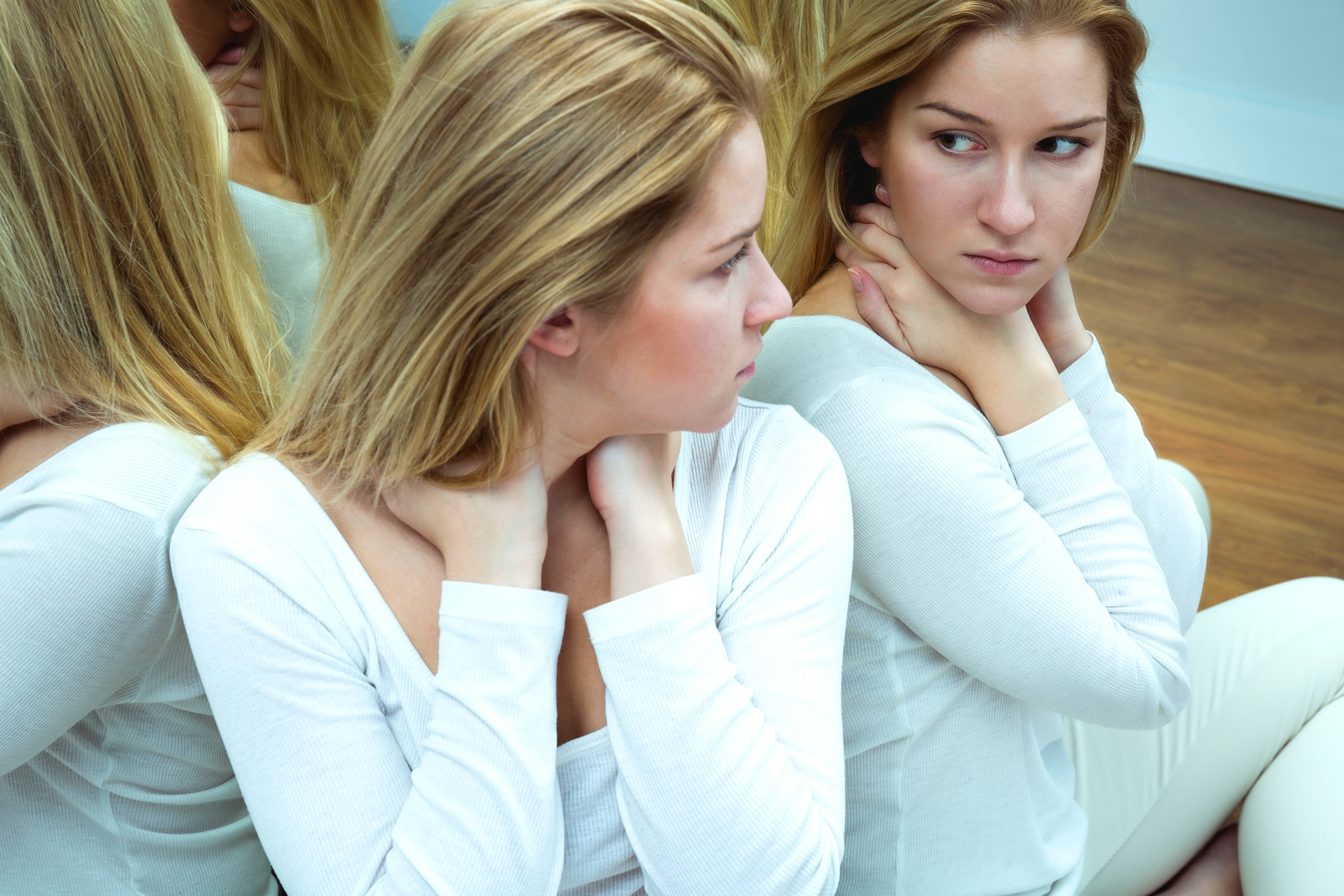 A woman with blonde hair, wearing a white long-sleeved shirt, is sitting in front of a mirror. She is looking at her reflection with a thoughtful expression, her hands clasped around her neck. Multiple reflections are visible around her.