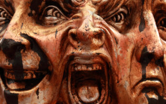 Close-up of a wooden sculpture featuring three expressive faces tightly placed next to each other. The left face has a sinister smile, the central face appears to be screaming in anger, and the right face shows sadness or despair. The sculpture is painted in earthy tones.