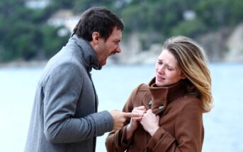 A man and woman stand outdoors near a body of water and trees. The man, wearing a gray coat, is shouting and pointing aggressively at the woman, who looks distressed and is holding her coat closed. The woman’s expression shows pain and fear.