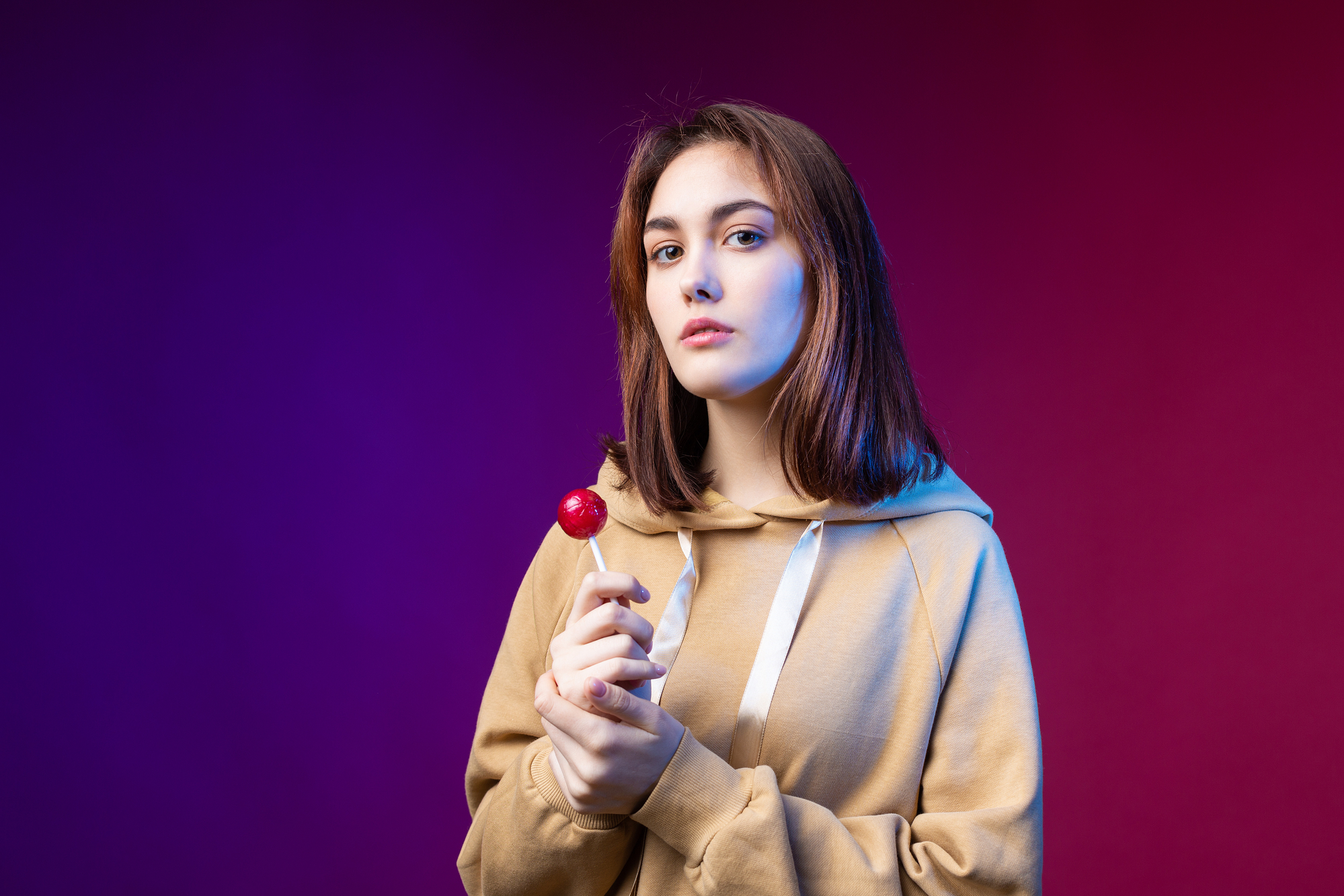 A young woman with shoulder-length brown hair, wearing a beige hoodie, stands against a gradient purple and red background. She holds a red lollipop in both hands, looking directly at the camera with a neutral expression.