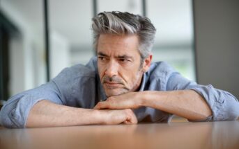 A middle-aged man with salt-and-pepper hair and a beard leans on a table, looking pensive. He is wearing a light blue button-up shirt and rests his chin on his forearm, appearing deep in thought. The background is out of focus, suggesting an indoor setting.