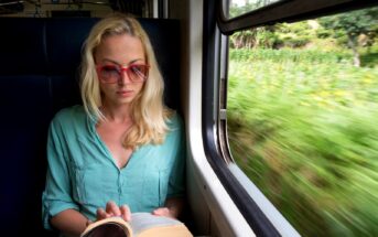 A woman with blonde hair and red-framed glasses is seated by a window in a train, reading a book. She is wearing a light teal shirt. The view outside the window shows green, blurred scenery, indicating the train is moving.