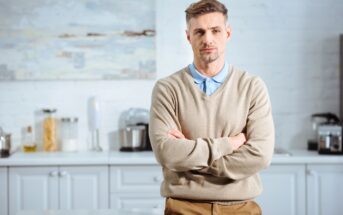 A man with short hair stands in a modern kitchen with his arms crossed. He is wearing a beige sweater over a light blue collared shirt and tan pants. The kitchen has white cabinets and various kitchen appliances on the counter.