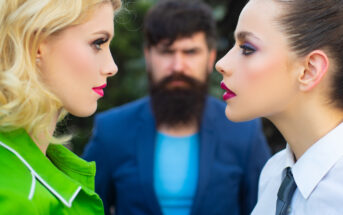Two women face each other with serious expressions, one with blonde hair in a green jacket and the other with dark hair in white attire. A bearded man in a blue suit stands in the background, looking at them with a stern expression. The background is blurred.