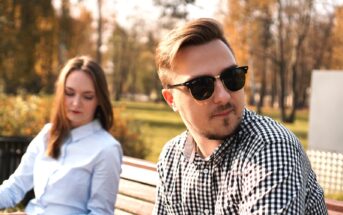 A man wearing sunglasses and a checkered shirt looks to the side while sitting on a bench in a park. A woman with long brown hair, wearing a blue button-up shirt, sits next to him looking down. The background features trees with autumn leaves.